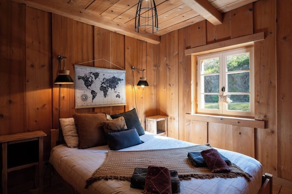 A wooden-paneled room with a bed, two wall-mounted lamps, a world map, and a window with a view of greenery.