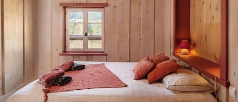 A bedroom with a wooden floor, a bed with a red blanket, two pillows, a window, and a bedside table with a lamp.