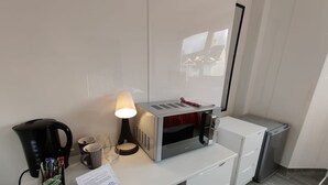 Quarto de hotel com uma escrivaninha branca, micro-ondas, chaleira, luminária e lixeira.
