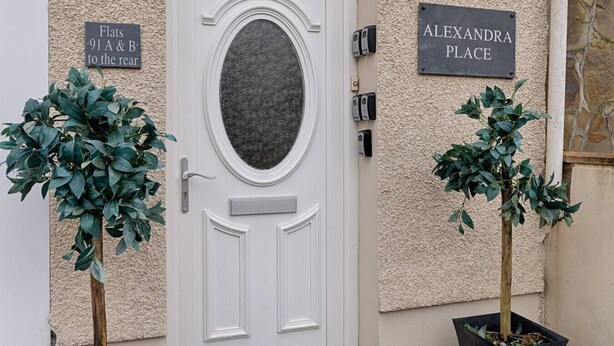 A white door with an oval glass panel, two signs indicating 'Flats 91 A & B to the rear' and 'Alexandra Place', and two potted plants beside the door. - 1 Bed Home • Parking • Great for Contractors (Gorseinon)