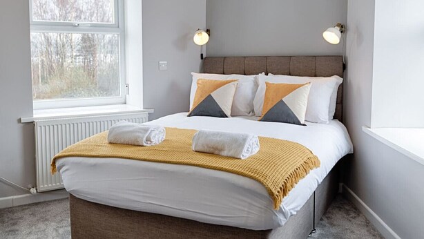A neatly made bed with a white and yellow blanket, two pillows, and two rolled towels. There is a window with a view of trees, a wall-mounted light fixture, and a radiator.