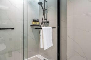 A glass-enclosed shower with a handheld showerhead, a towel rack with a white towel, and two bottles on a shelf.