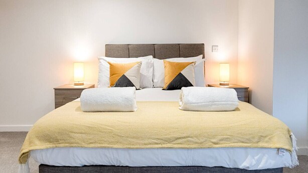 A neatly made bed with a headboard, two bedside tables with lamps, and a yellow throw blanket. - Contractor Base • Kitchen • Parking Included (Plasmarl)
