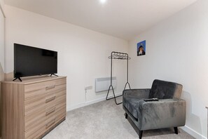 A room with a flat-screen TV on a wooden dresser, a grey sofa, a floor lamp, and a radiator.