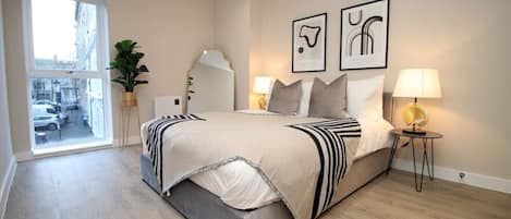 A modern bedroom with a large bed, bedside lamps, a window with a view, and two framed artworks on the wall.