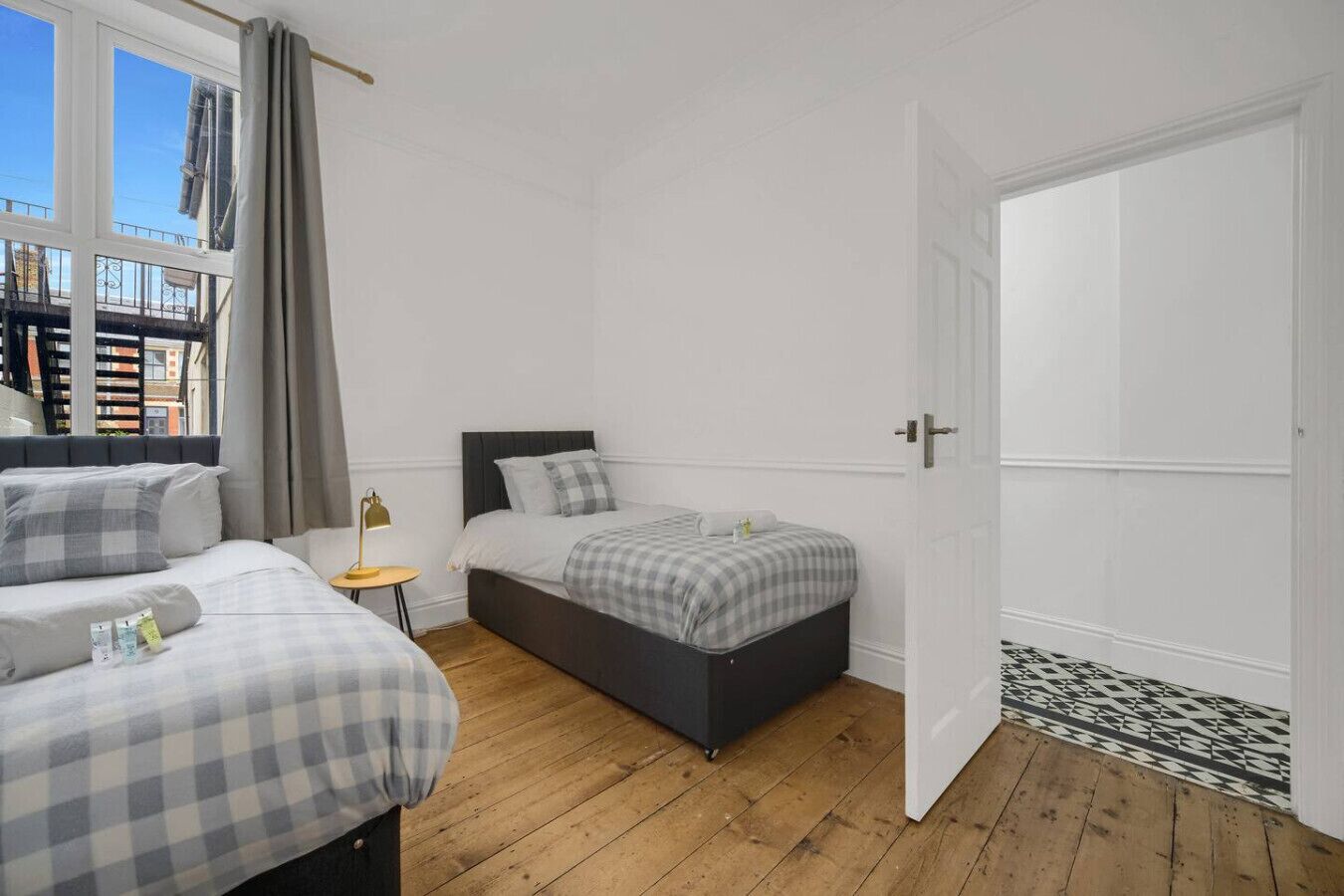 A bedroom with two beds, a bedside table, a lamp, and a view of buildings outside.
