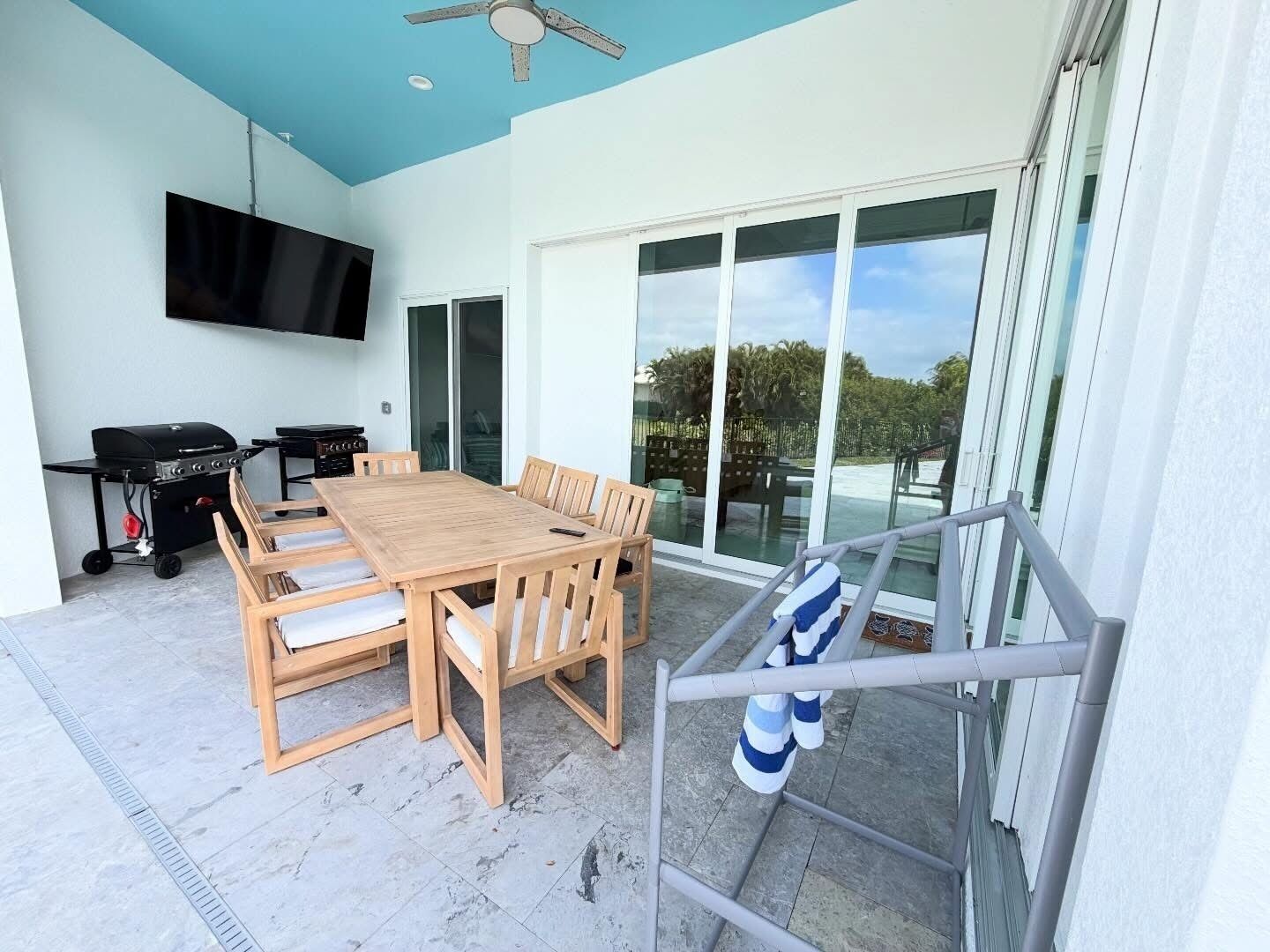 An outdoor patio with a wooden table and chairs, a grill, and a TV mounted on the wall.