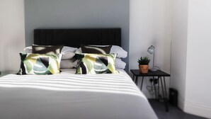 Una cama bien hecha con ropa de cama a rayas, un cabecero oscuro y almohadas estampadas. Se observa una mesita de noche con una lámpara y una planta en maceta.