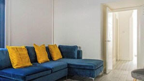 Una sala de estar con un sofá azul, tres almohadas amarillas y una puerta que da a otra habitación.