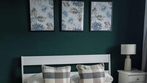 En säng med en vit sänggavel, två kuddar, ett nattduksbord med en lampa och tre inramade konstverk med blommotiv på väggen.