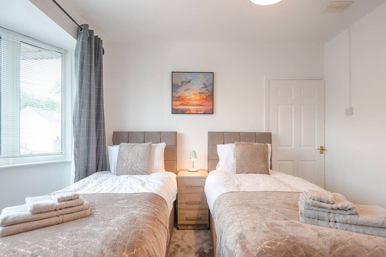 A double room with two beds, a nightstand with a lamp, a painting on the wall, and a window with curtains.