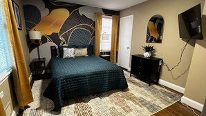 Ein Schlafzimmer mit Bett, Nachttisch, Kommode, Wandfernseher und einer Tür.