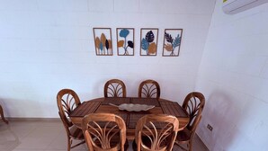 A round wooden dining table with six chairs, white tiled floor, and four framed wall art pieces above the table. - Your Home way From Home (San Salvador)