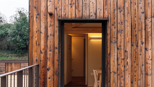 Un bâtiment à l’extérieur en bois, avec une porte bien visible donnant sur un espace intérieur où se trouvent une chaise et une table.
