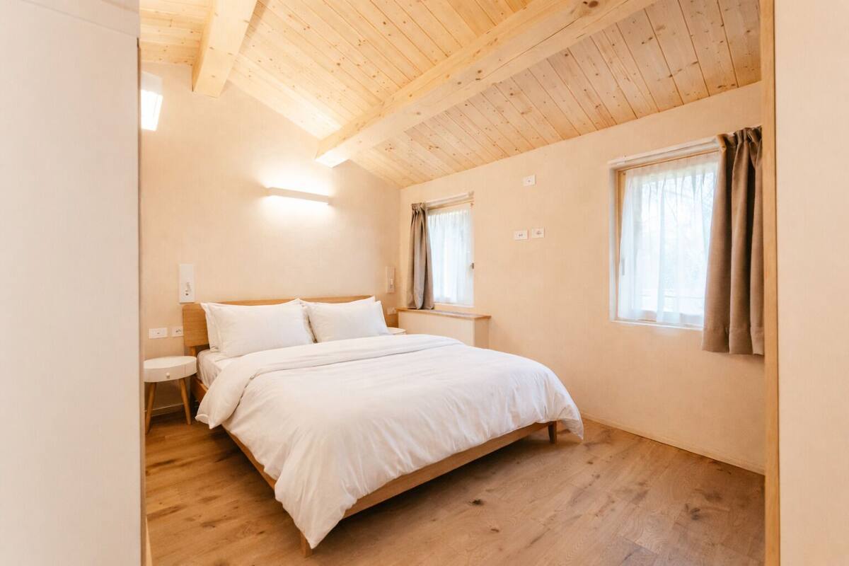Un dormitorio con techo de madera, una cama con ropa blanca, una mesita de noche, una ventana con cortinas y una pequeña mesa redonda.