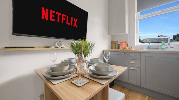 Una cucina moderna con un tavolo da pranzo per quattro persone, una TV a parete che mostra 'NETFLIX' e una finestra con vista su un cantiere.
