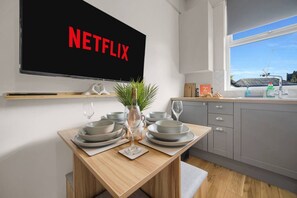 A modern kitchen with a dining table set for four, a TV mounted on the wall displaying 'NETFLIX,' and a window with a view of a construction site. - 2 Streets from Centre | Free Parking | Fast WIFI (Cardiff)
