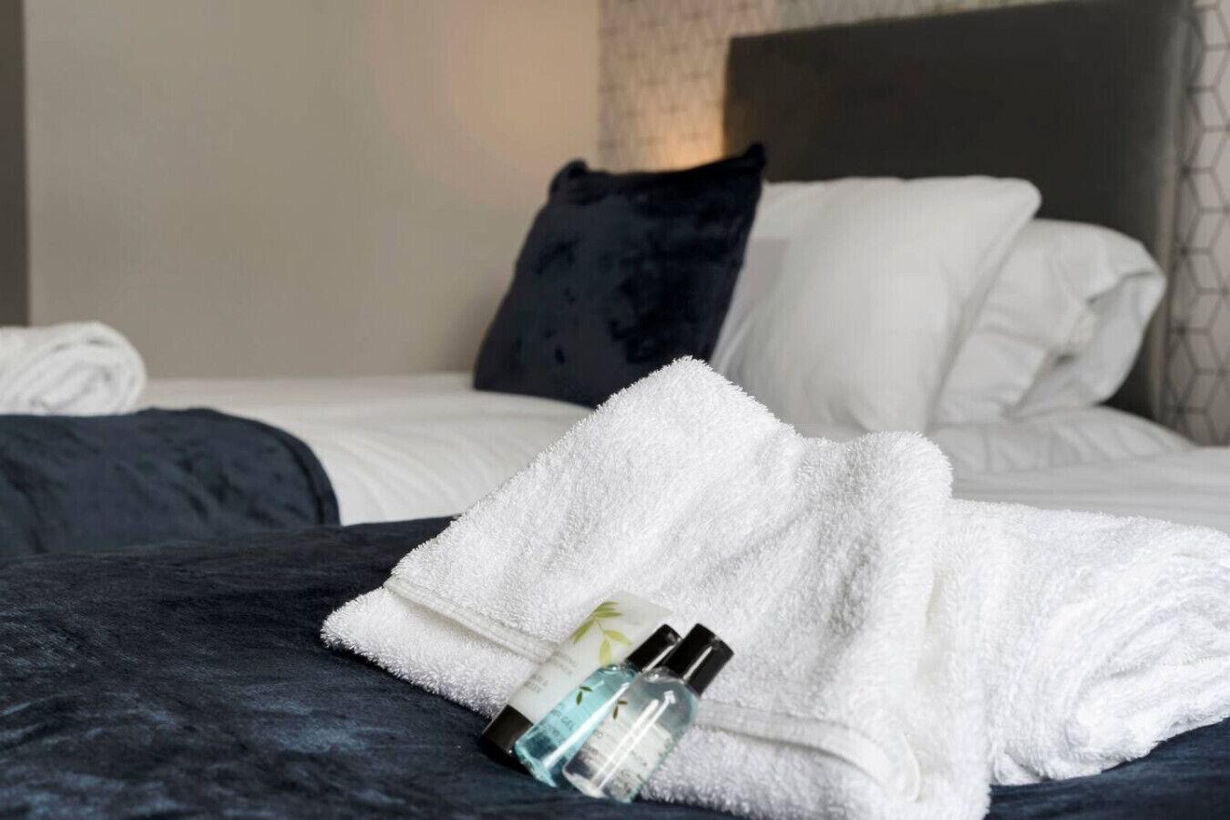 A neatly made bed with a white towel, a dark blue blanket, and two bottles of lotion.