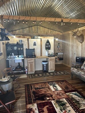 Ein rustikales Interieur mit Holzwänden, einem an der Wand aufgehängten Bärenkopf, einem gegenüberliegenden Hirschkopf, einer Küchenzeile mit Spüle und Arbeitsplatte sowie einer Sitzecke mit Sofa und Sesseln.