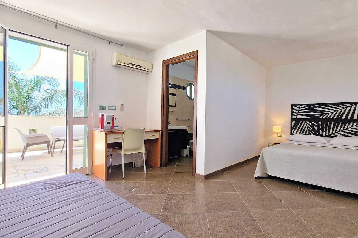 A hotel room with a bed, a desk with a chair, a bathroom, and a view of palm trees.