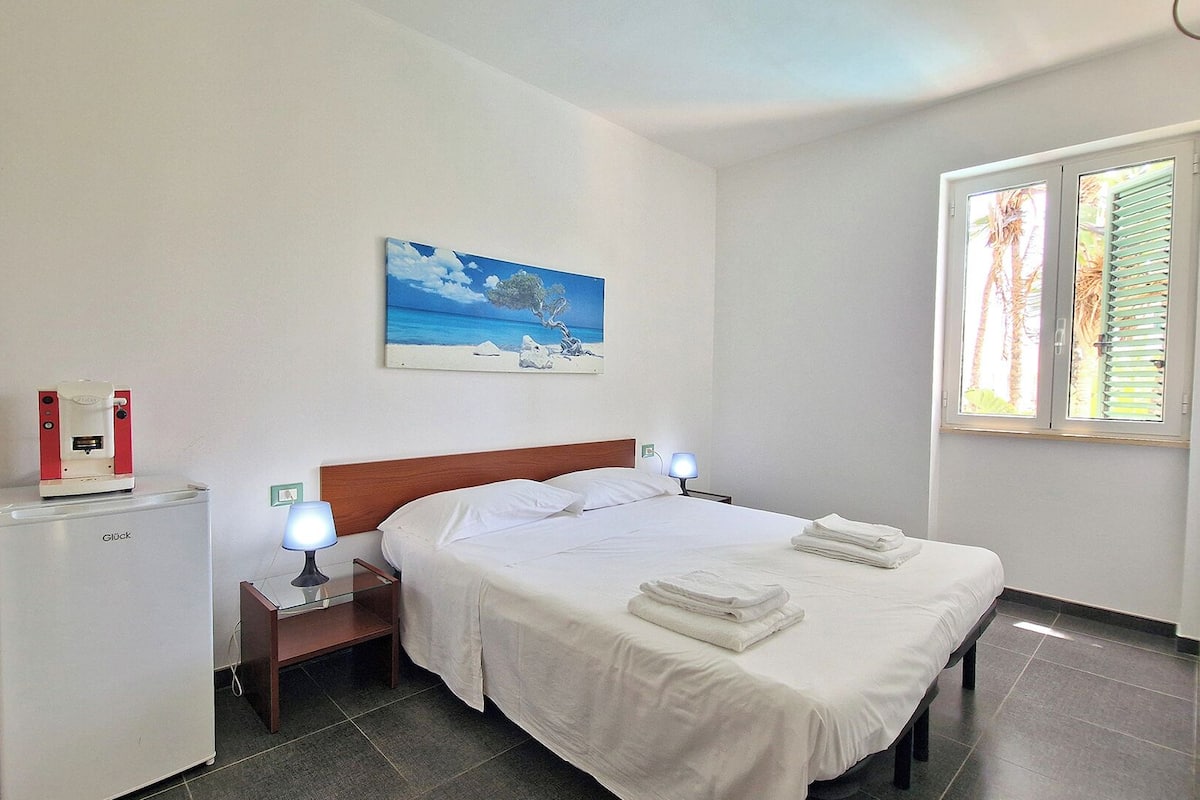 A hotel room with a bed, bedside lamps, a small table, a painting, and a window with blinds.