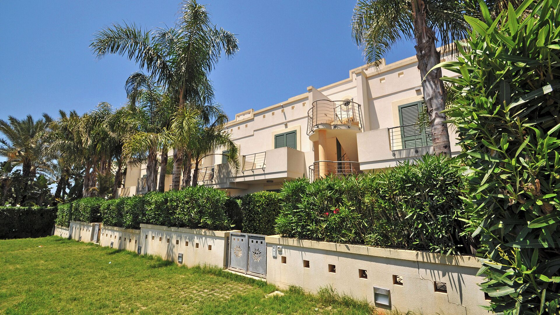A multi-story residential building with balconies, a concrete wall with small windows, and lush greenery.