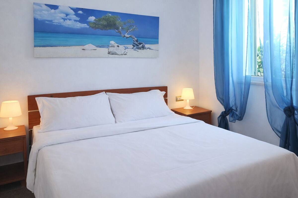 A hotel room with a bed, two bedside tables, a painting of a beach scene, and blue curtains.