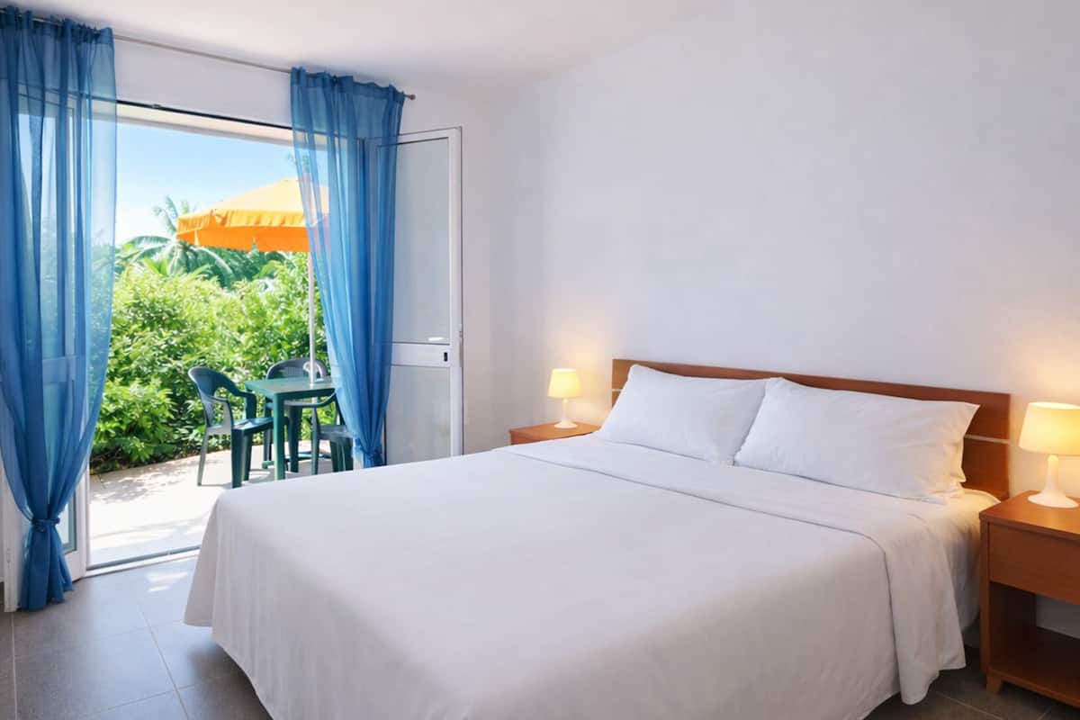 A hotel room with a bed, two bedside lamps, a wooden nightstand, a window with blue curtains, and a view of greenery outside.