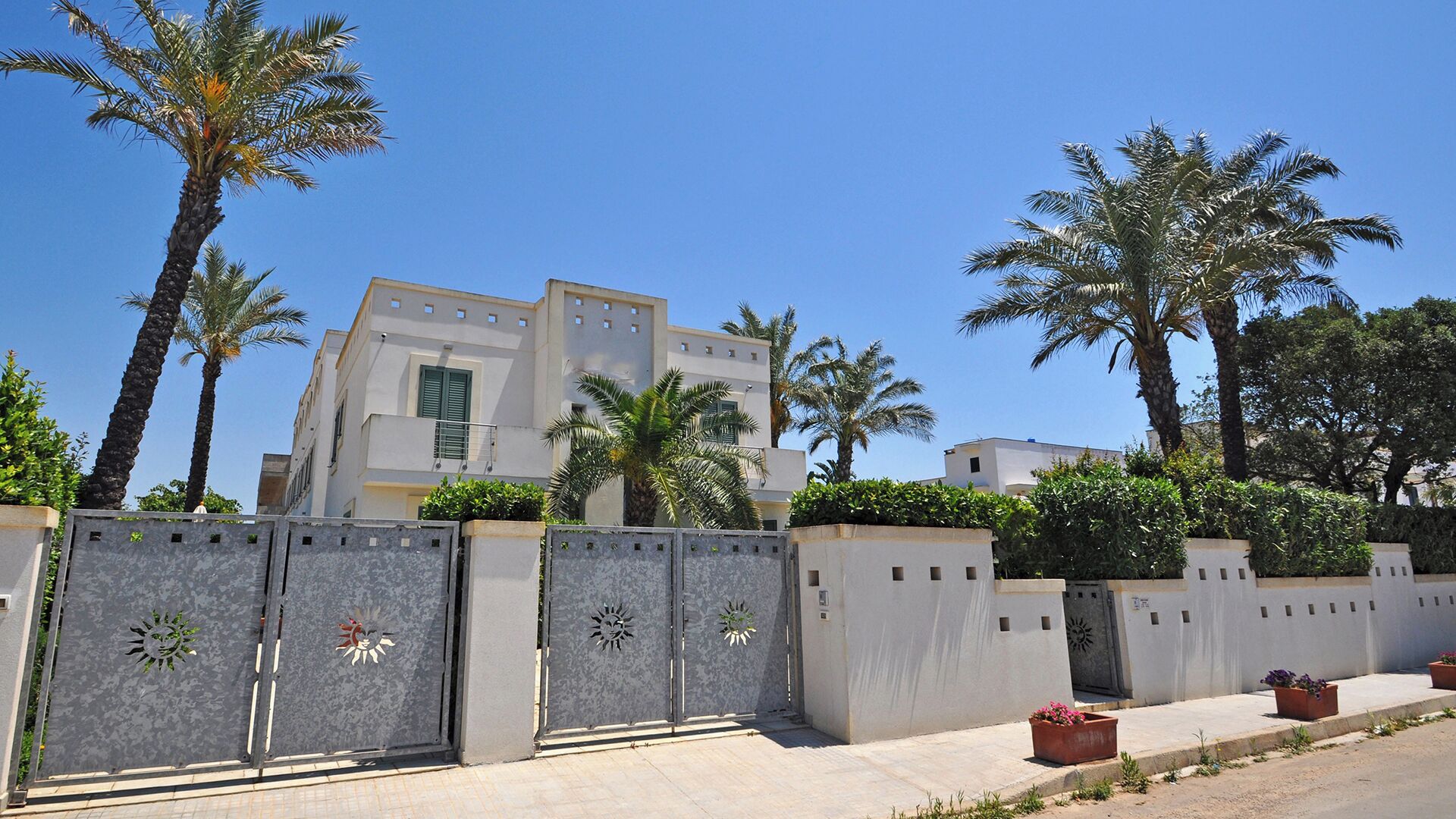 A modern building with a white facade, surrounded by palm trees and a gated entrance with decorative metalwork.