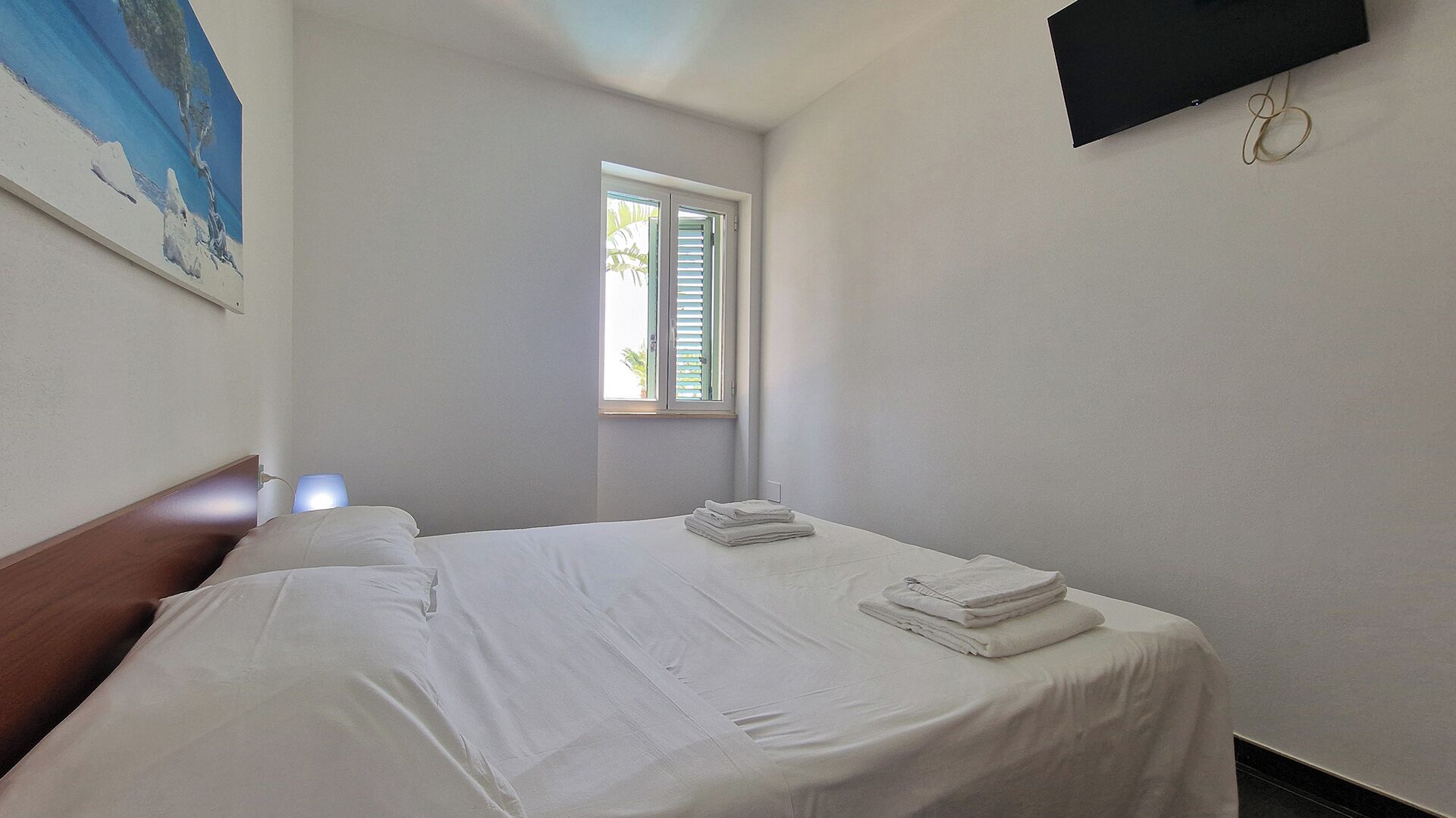 A bedroom with a bed, a window, a painting on the wall, and a TV mounted on the wall.