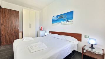 Una camera d'albergo con un letto, un quadro raffigurante una spiaggia, un comodino con una lampada e un armadio in legno.