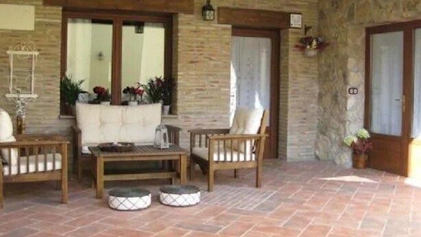 Un patio couvert avec des murs en pierre, un plafond en bois, un canapé blanc, une table en bois et deux bancs rembourrés.