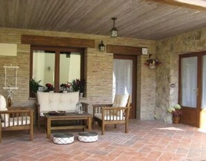 Ein überdachter Patio-Bereich mit Steinwänden, Holzboden und einer weißen Couch, einem Holztisch und zwei gepolsterten Bänken.