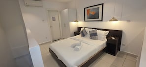 A neatly made bed with pillows, a bedside table, a framed picture on the wall, and a door in the background. - EBS Properties At Embassy Garden (Accra)