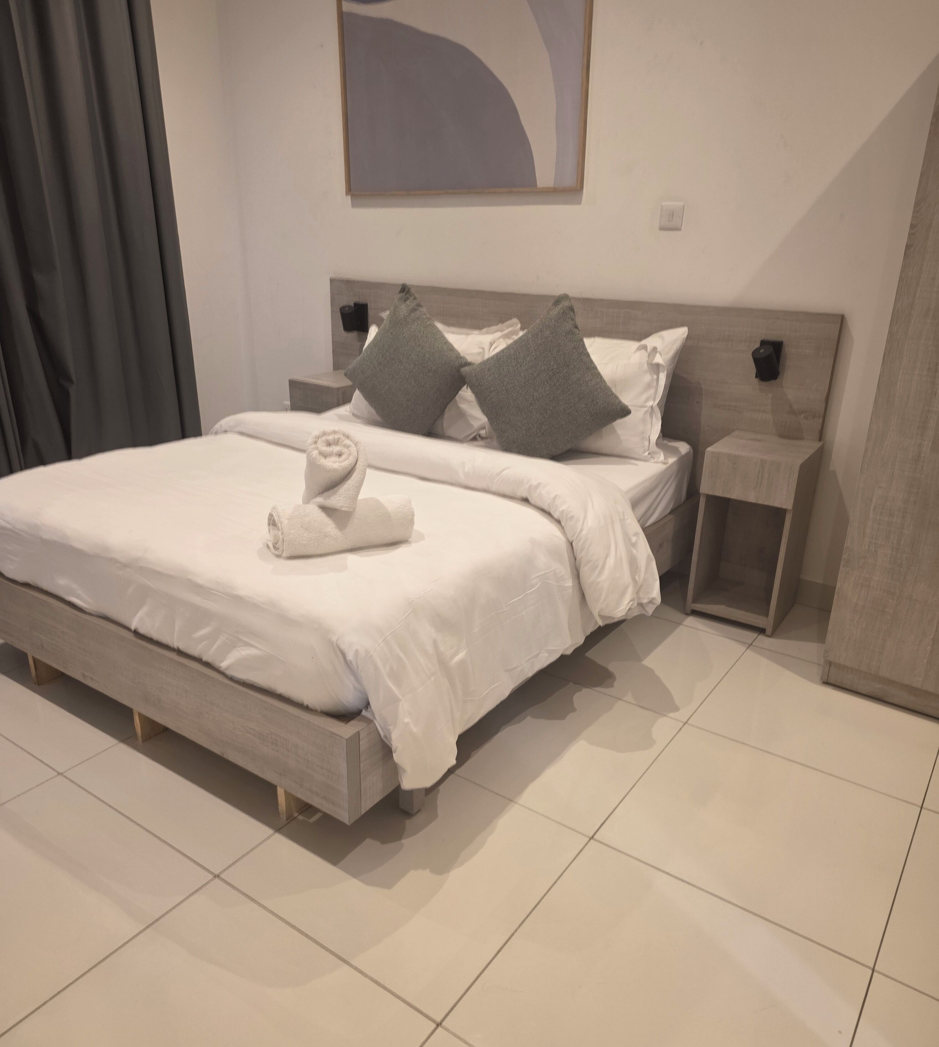 A neatly made bed with a towel, a wooden headboard, a nightstand, and a wall-mounted light fixture.