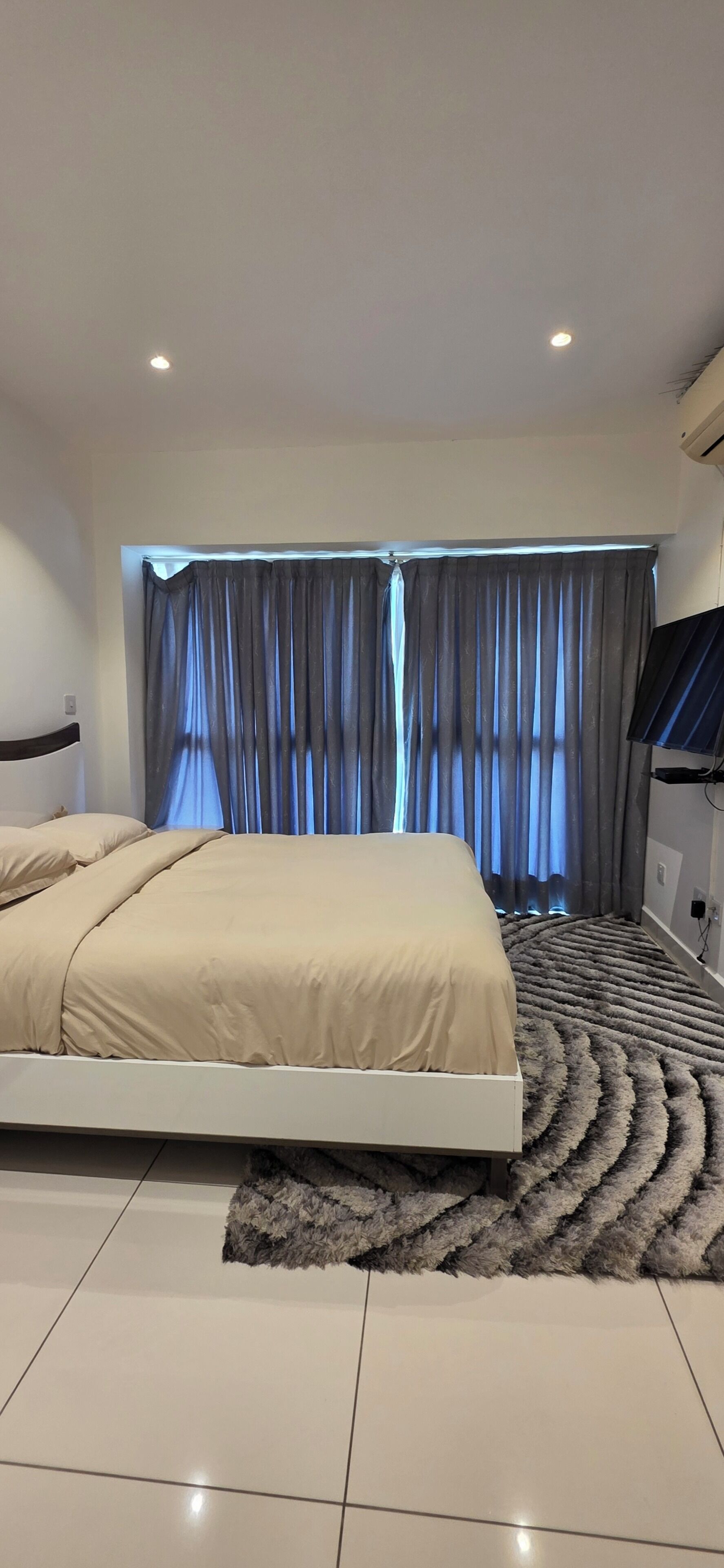 Een moderne slaapkamer met een bed, gordijnen, een televisie en een kamerkleed met een patroon.