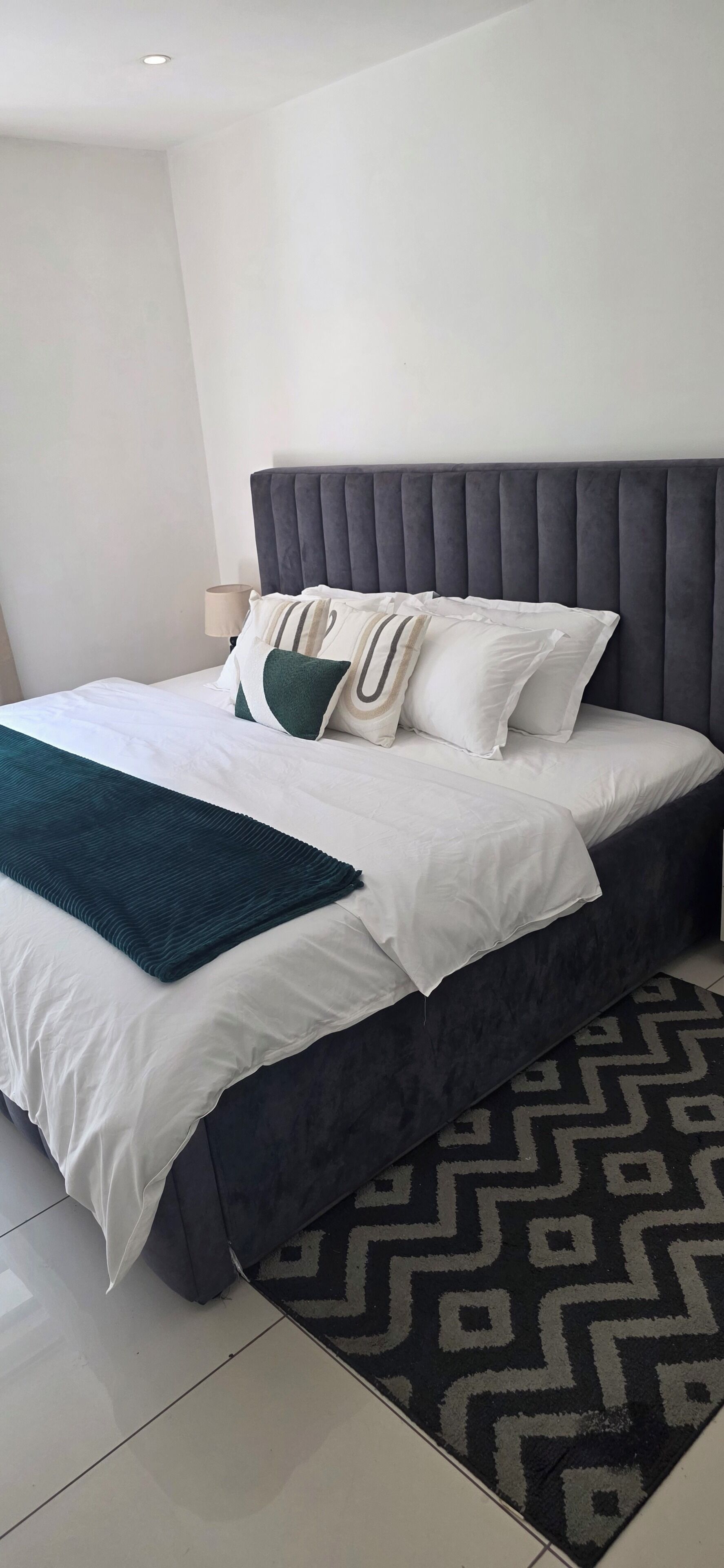 Een netjes opgemaakt bed met een grijs bekleed hoofdbord, wit beddengoed en een groene plaid.