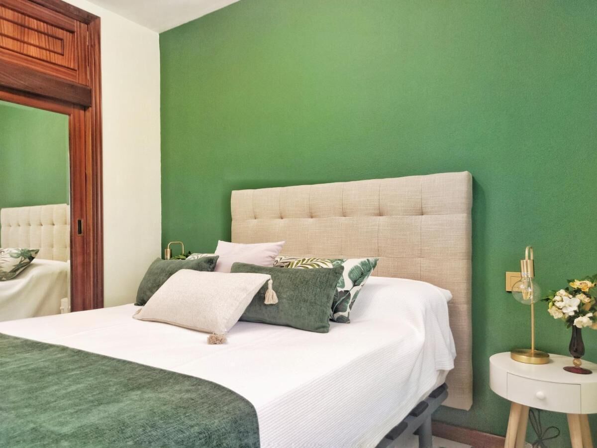 Een slaapkamer met een groene wand, een gestoffeerd hoofdbord, een witte spreideken en een nachtkastje met een lamp.