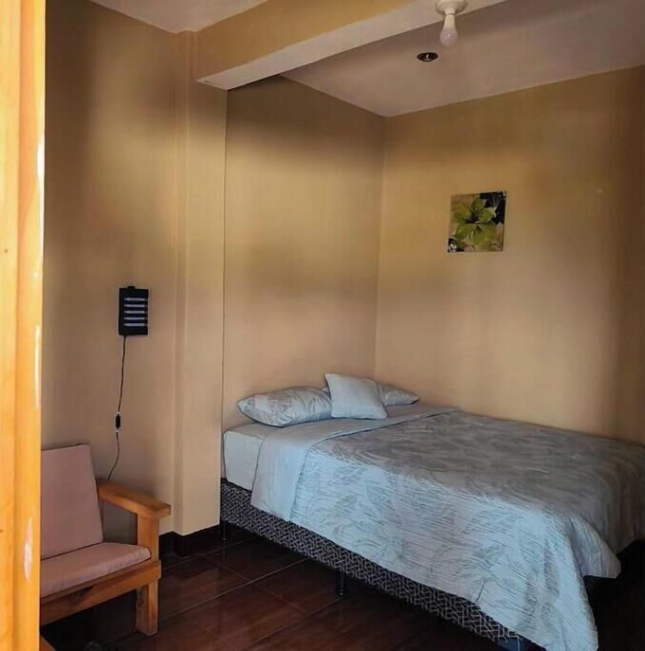 Une petite chambre avec un lit simple, une chaise en bois, une prise de courant et un luminaire au plafond.