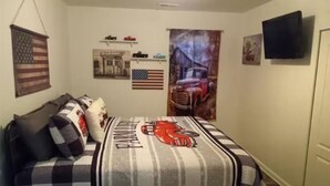 A bedroom with a checkered bedspread, an American flag, a vintage car poster, and a television on the wall. - Creekside Garage Getaway  (Robbinsville)