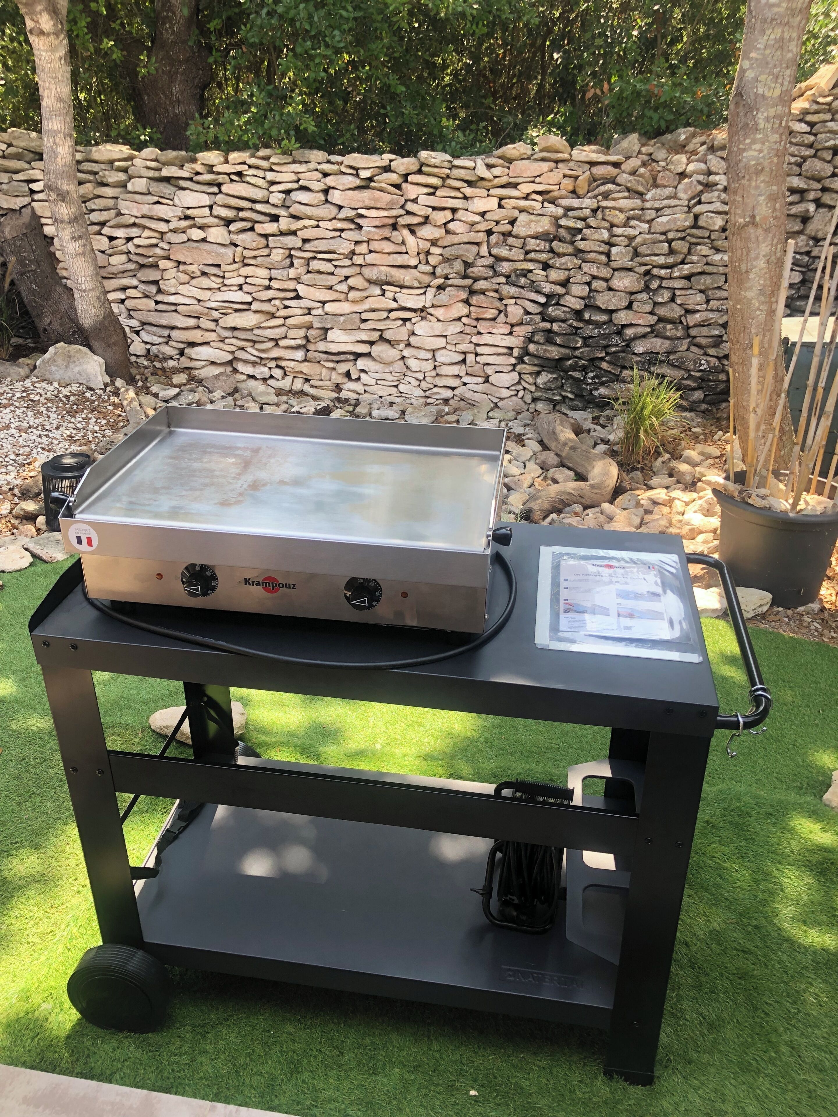 A portable grill on a cart with a stone wall and trees in the background.