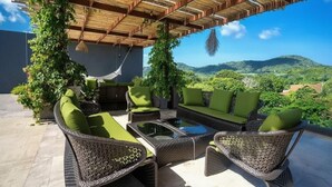 Un área de estar al aire libre con techo, mobiliario de mimbre y cojines verdes, con vistas a un paisaje montañoso.