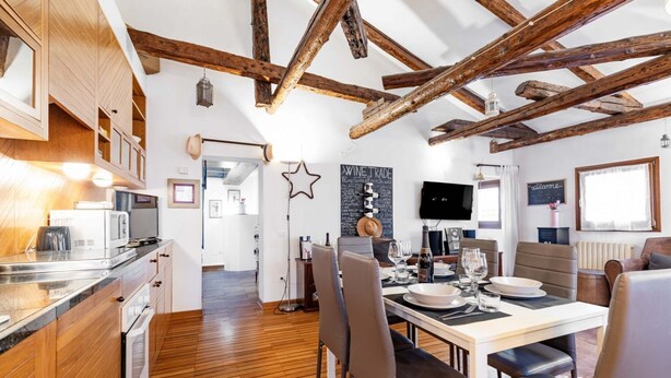 Una cocina moderna con vigas de madera, un comedor con una mesa dispuesta para dos personas y una sala de estar con televisión.