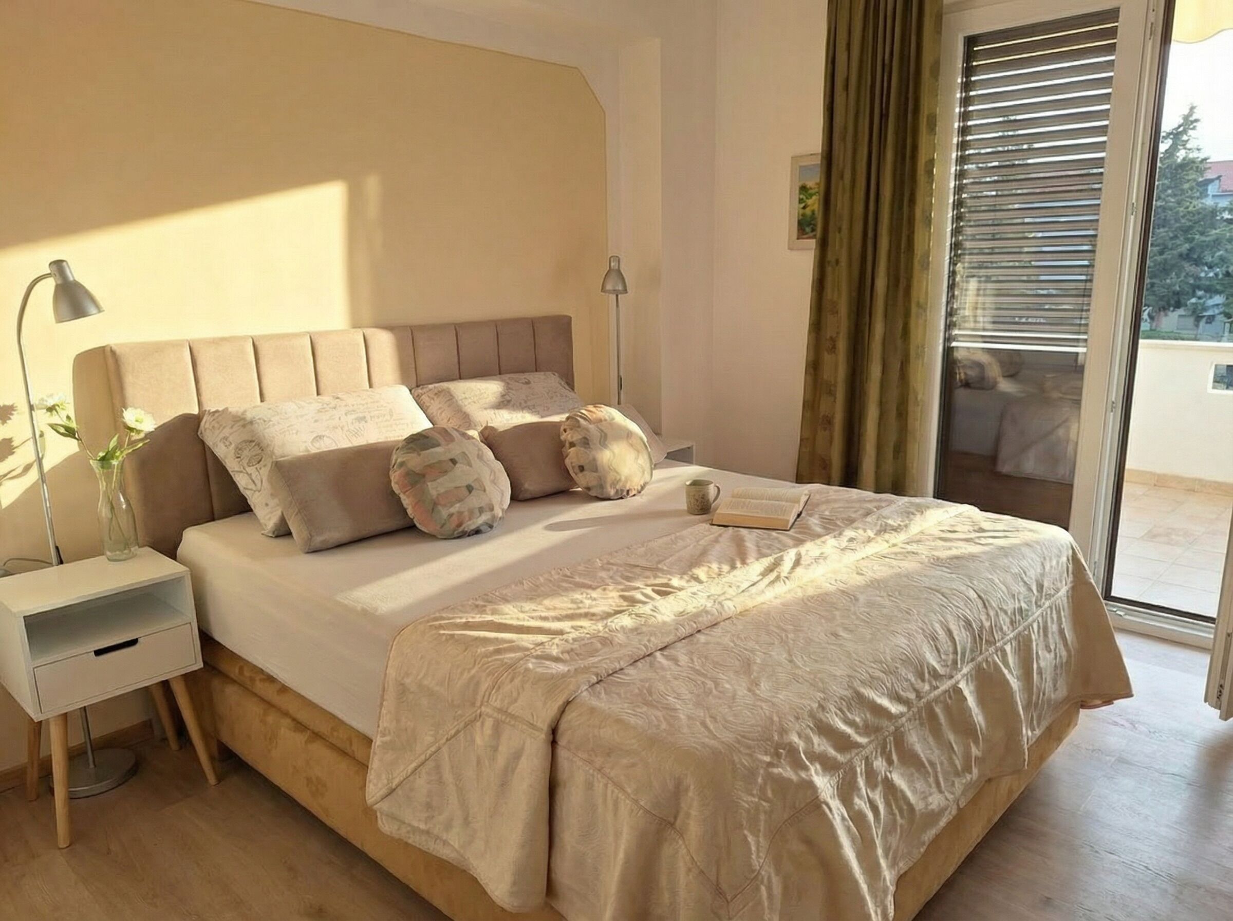 Un dormitorio con cama, mesitas de noche, lámpara, ventana con cortinas y vistas al exterior.