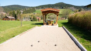 Una cancha de croquet con un pabellón de madera, un camino de grava y un paisaje rural con casas y colinas al fondo.