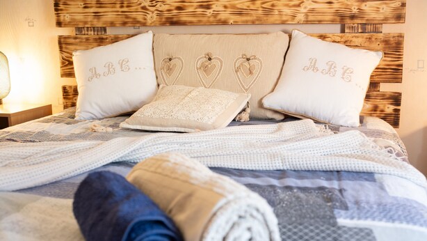Una cama bien hecha con toallas enrolladas, una colcha estampada y almohadones con forma de corazón e iniciales.