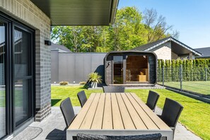 Eine moderne Außenpatio mit Holztisch und -stühlen, einer überdachten Sitzgruppe und Blick auf ein Haus mit großem Fenster und umzäunter Garten.