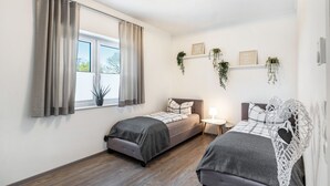 Un dormitorio con dos camas, una ventana con cortinas, una mesita de noche con lámpara y estantes en la pared con plantas.