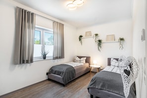Ein Schlafzimmer mit zwei Betten, einem Fenster mit Vorhängen, einem Nachttisch mit Lampe sowie an der Wand befestigten Regalen mit Pflanzen.