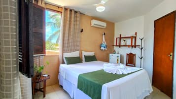Un dormitorio con cama, aire acondicionado, una mesita con una planta y vistas a palmeras fuera.